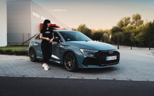 Audi RS3 SB szemből, naplementében, egy fekete hajú nővel az előtérben, szemből.
