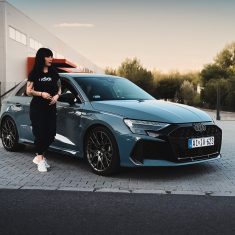 Audi RS3 SB szemből, naplementében, egy fekete hajú nővel az előtérben, szemből.