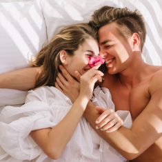 Cheerful young couple in love lying in white bed, enjoying honeymoon in sunny morning. Smiling handsome man with bronze skin embracing his magnificent girlfriend in vintage blouse resting on pillows.