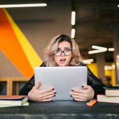 portrait of young pretty woman with socked face expression, sitting at table working on laptop in co-working office, wearing glasses, stress at work, funny emotion, student in class room, frustration