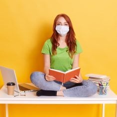 Young student in medical mask studying at home while quarantine, bored of distance learning, sitting with crossed legs on white table with book in hands.