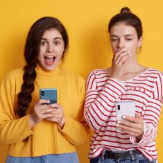 Two friends with phones reading shocking news in social network, lady with pigtail looking at camera with big eyes and yelling something, female in striped shirt covering her mouth with palm.