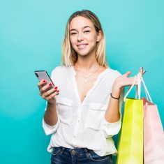 Portrait of a happy young woman holding shopping bags and mobile phone isolated on a mint background