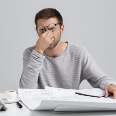 Stess, overwork and deadline. Tired unshaven young architect freelancer massaging nose bridge , having headache while working in big architectural project at home office, feeling exhausted and sleepy