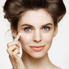 Young beautiful brunette girl in hair curlers smiling looking at camera creaming face over white background. Facial treatment. Beauty health and cosmetology.