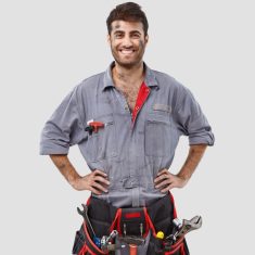 Buoyant successful handyman wears grey overalls, tool belt, stands with hands on waist over white background has shining smile. Content industrial worker rejoices coming weekend after hard working day
