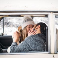 A Valentine couple sitting inside a snow retro car and kissing in a forest. Romantic Beauty girl in a hat and handsome man in a grey pullover. Horisontal picture