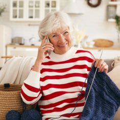 Aged people, retirement, leisure and modern technology concept. Beautiful happy grandma with gray hair talking to her granddaughter on mobile phone while knitting scarf on couch in living room