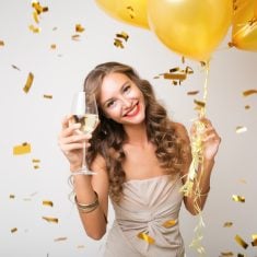 attractive young stylish woman celebrating new year, drinking champagne holding air balloons, golden confetti flying, smiling happy, white background, isolated, wearing party dress