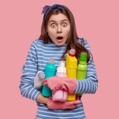 Photo of emotional European housemaid opens mouth widely, stares in stupor, carries bottles of cleaning supplies, cares of sanitary and hygiene, wears striped jumper, isolated over pink background